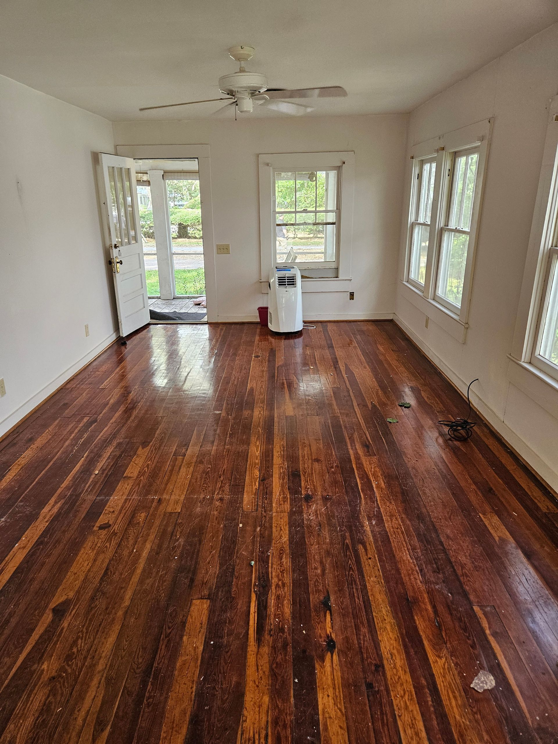 Empty room with hardwood floors and white walls, windows on the right, door on the left, and an air purifier in the center.