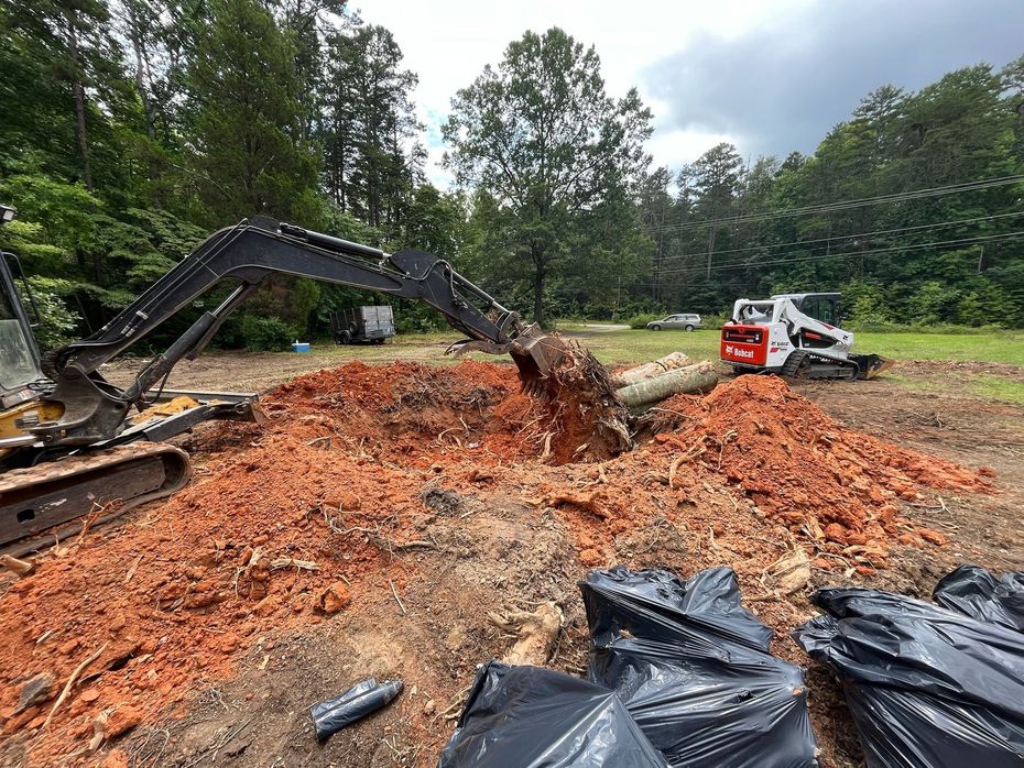 An excavator digs into a pile of red dirt, removing a tree trunk in Greensboro, NC