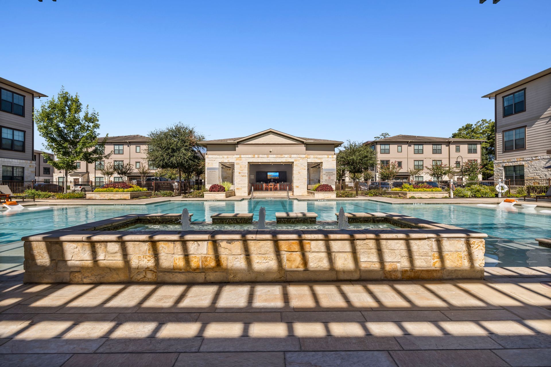 Pool with fountain, surrounded by apartment buildings under a blue sky.