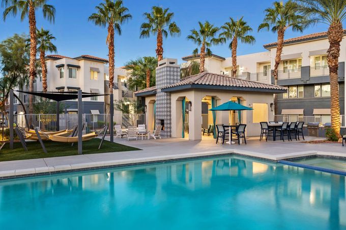Poolside scene with palm trees, a lounge area, and apartment buildings in the background.