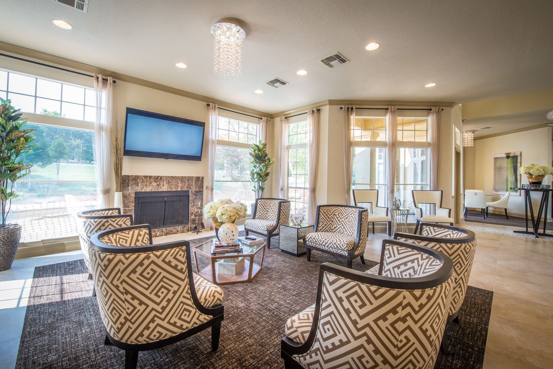 Lounge with patterned armchairs around a coffee table, fireplace, TV, and large windows.