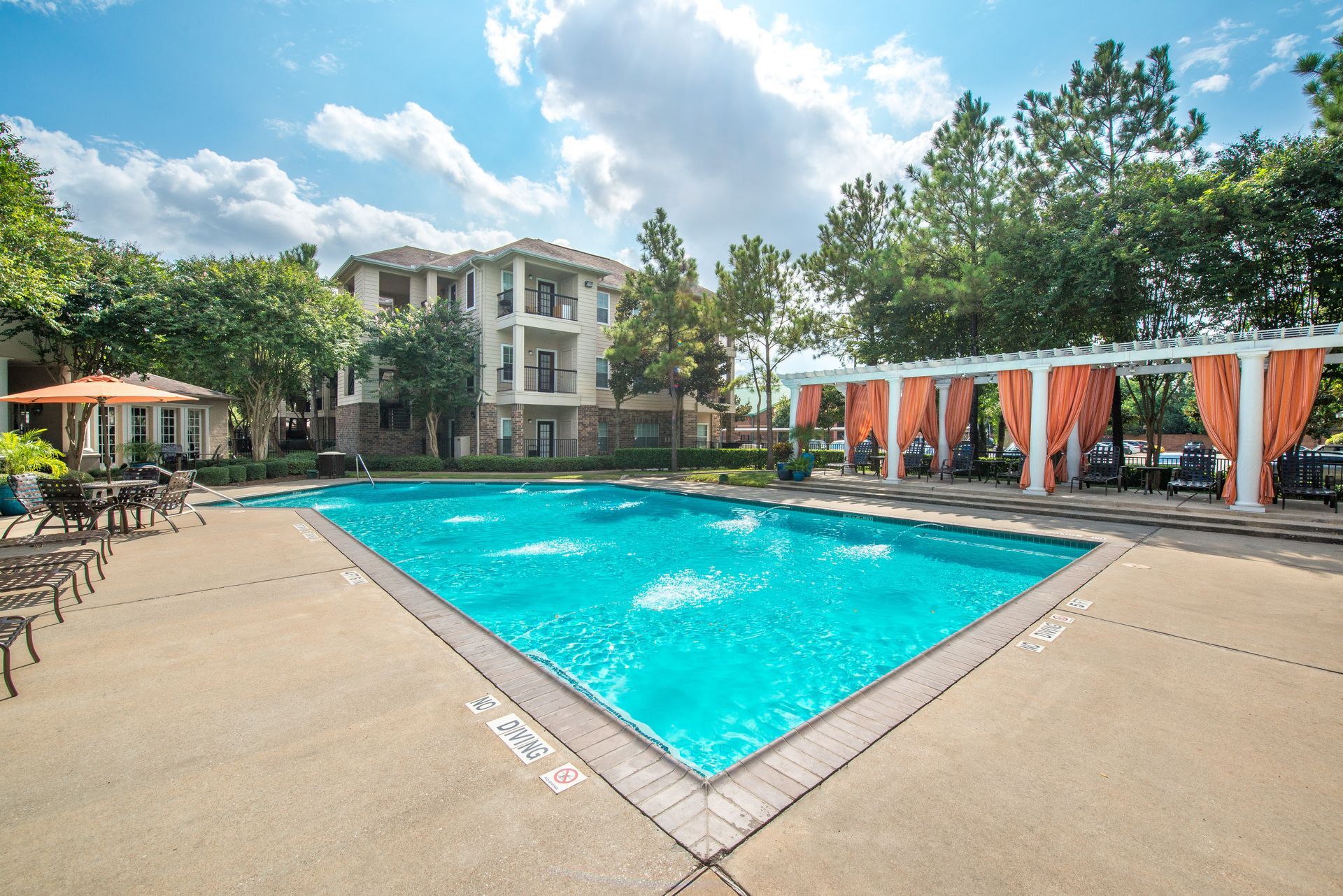 Swimming pool with fountains, beige patio, cabana with orange curtains, and apartment building in the background.