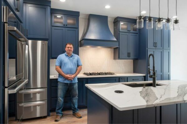 Man stands in a modern kitchen with navy cabinets, stainless steel appliances, and a white island.