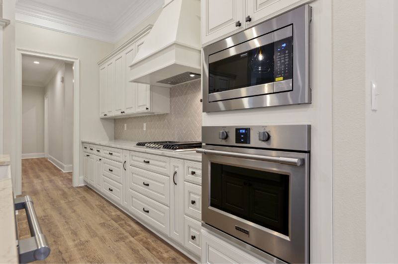 White kitchen with stainless steel appliances, wooden floors, and a hallway.