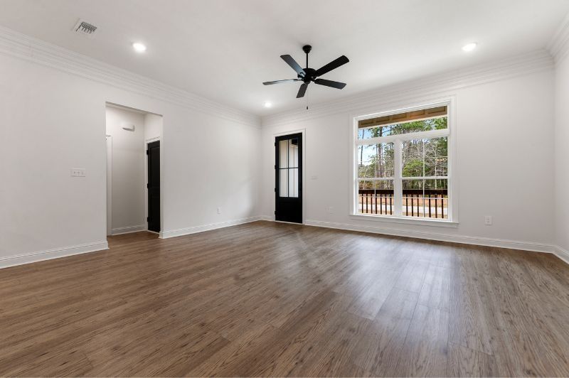 Empty, bright room with hardwood-look floor. White walls, black door, large window with view. Ceiling fan.