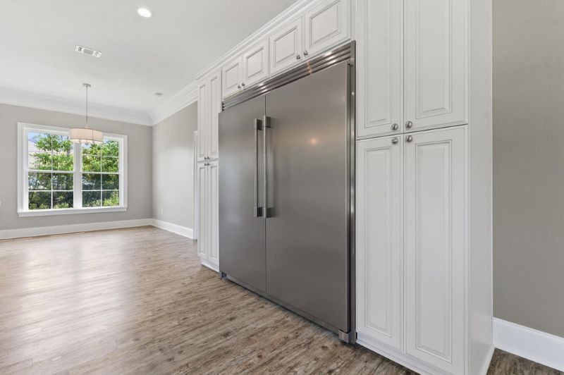 Stainless steel refrigerator in white kitchen cabinets. Window and wood floor in the background.