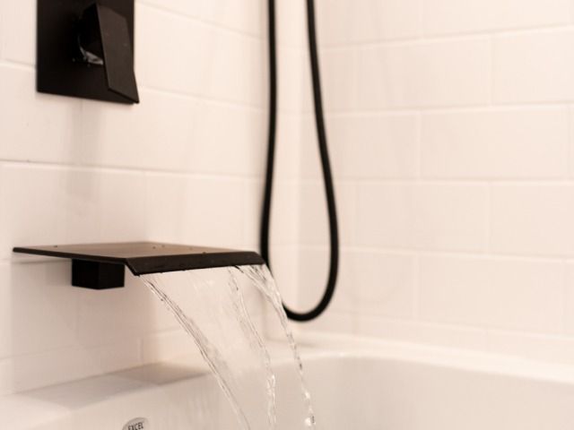 Black waterfall faucet pouring water into a white bathtub with white subway tile background.