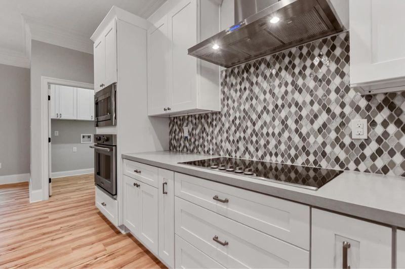 White kitchen with gray countertop, black and gray tiled backsplash, stainless steel range hood.