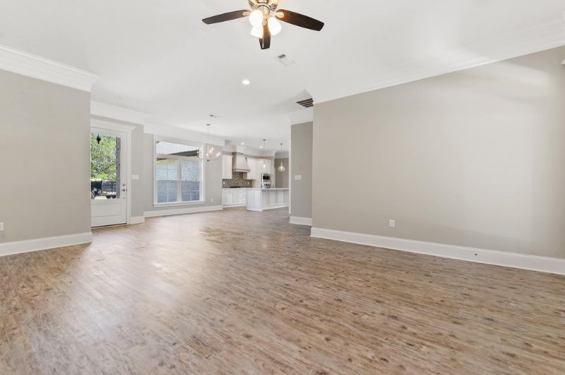 Empty living room with neutral walls, wood-look floors, ceiling fan, and a view into the kitchen.