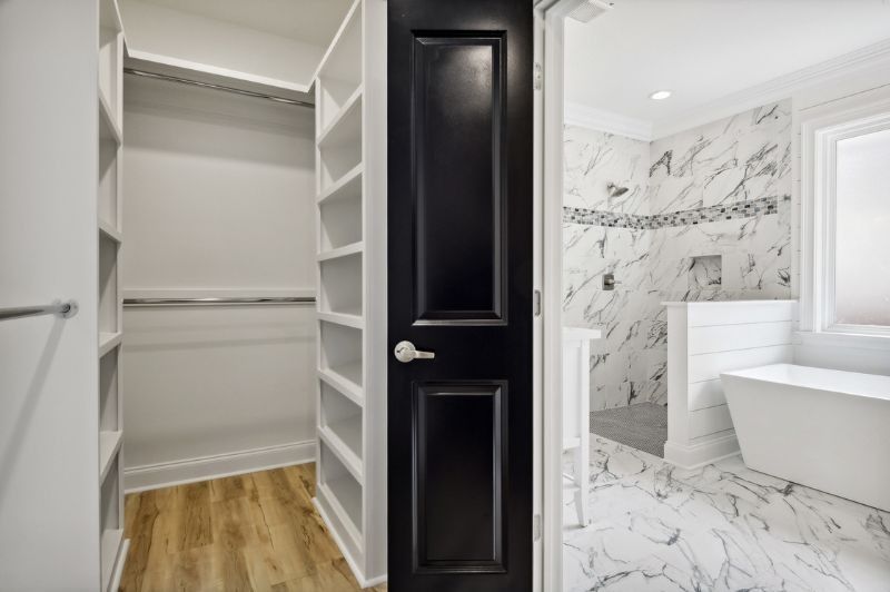 Walk-in closet with shelves and a black door leading to a modern bathroom with a marble shower and bathtub.
