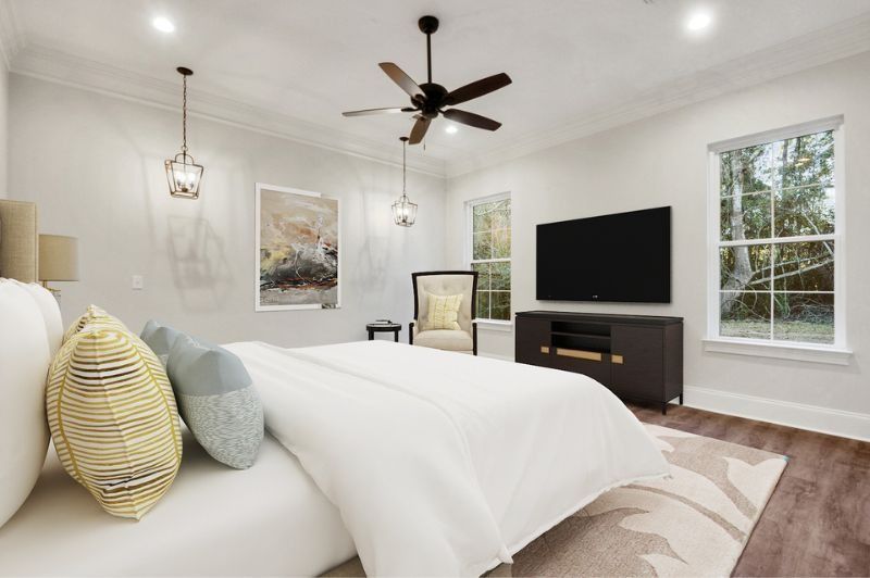 Bedroom with bed, TV, art, chair, windows, and a ceiling fan; neutral colors.