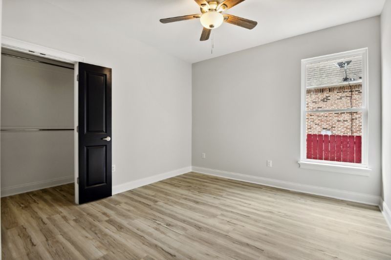 Empty bedroom with gray walls, wood floors, black closet door, and a window with red exterior.