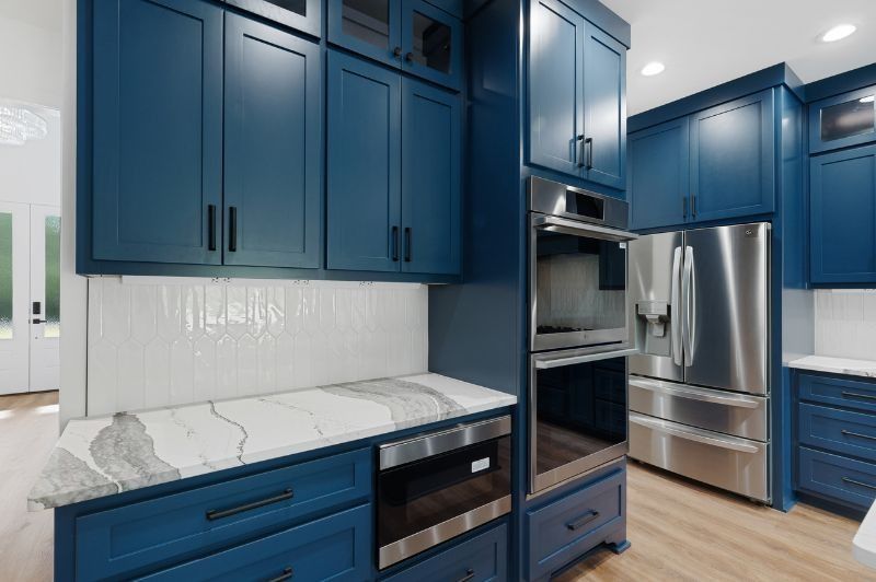 Blue kitchen cabinets, stainless steel appliances, white backsplash, marble countertop, and wood floor.