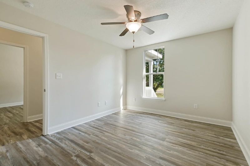 Empty room with light gray walls, wood-look flooring, and ceiling fan; window and door visible.