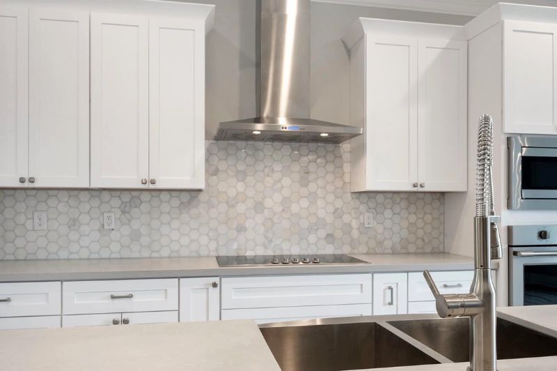 White kitchen with hexagon tile backsplash, stainless steel range hood, and white cabinets.