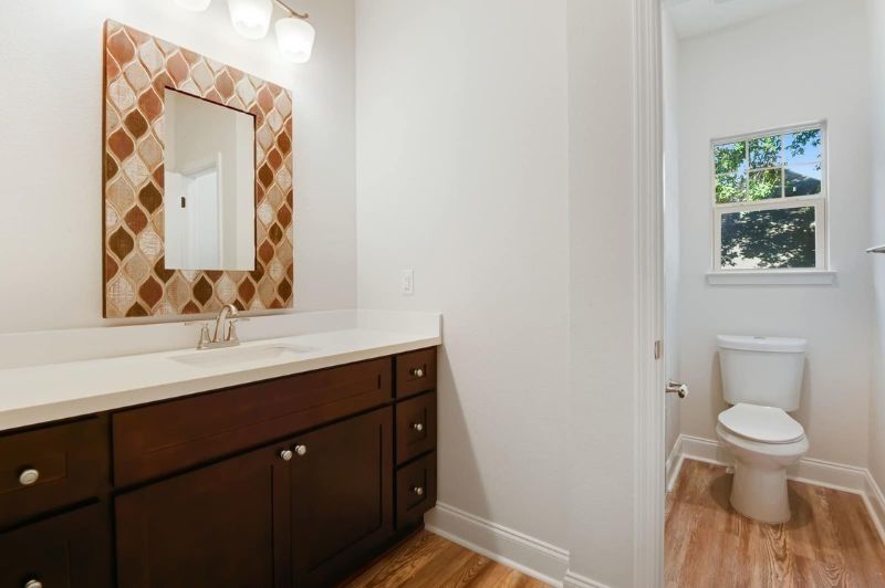 Bathroom with dark wooden vanity, patterned mirror, toilet, and window.