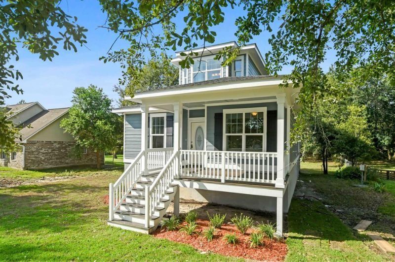 Blue cottage with white porch and stairs, green lawn, and surrounding trees.