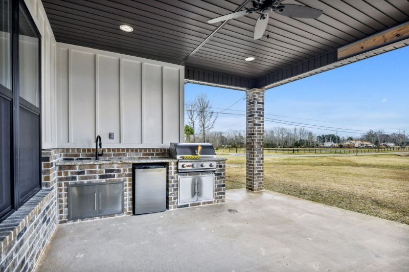 Outdoor kitchen with grill, sink, and fridge, under a covered patio with a view of a grassy field.