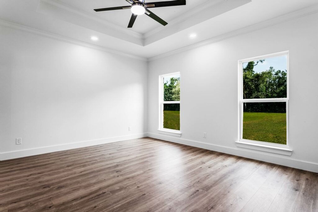 Empty room with hardwood floor, white walls, two windows overlooking a green yard, and a ceiling fan.