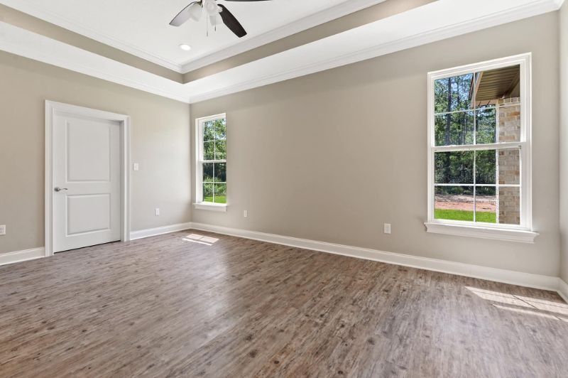 Empty bedroom with beige walls, white trim, and wood-look flooring; two windows and a closed door.