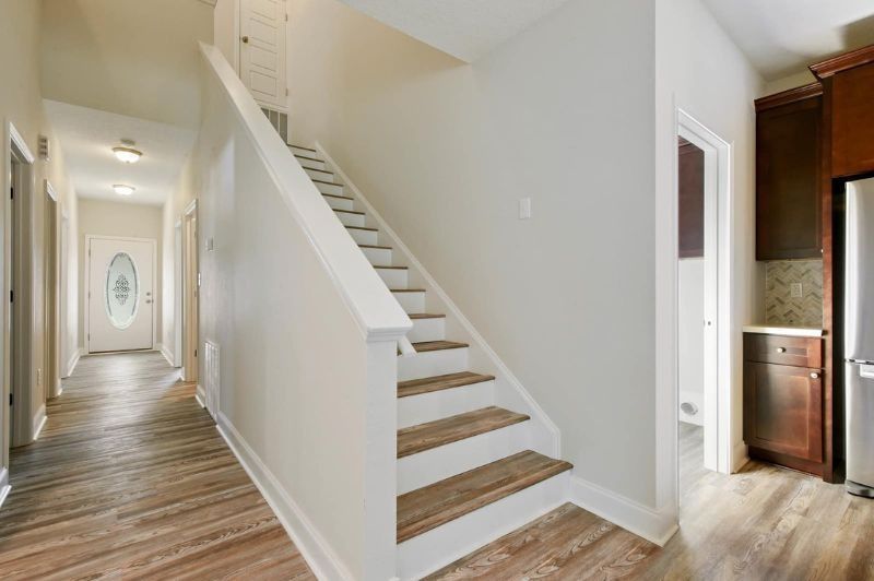 Hallway with staircase and kitchen visible. Light wood floors, white walls, wooden stairs.