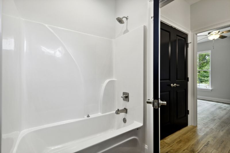 White bathtub and shower surround in a bathroom with a black door and wood floors.