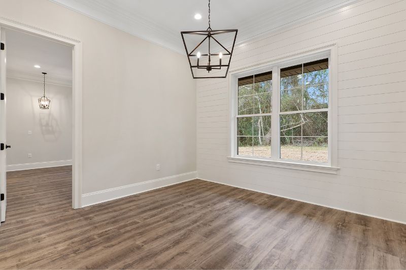 Empty room with wood-look floor and white shiplap wall. A chandelier hangs above, and a window looks outside.