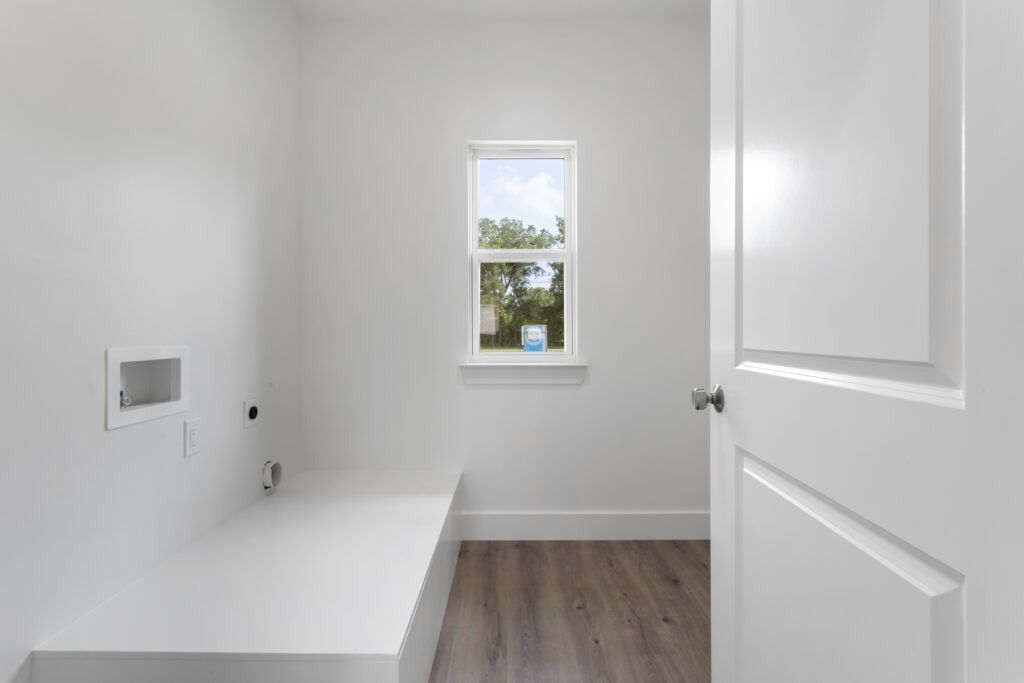 White laundry room with a window, door, and utility hookups. Light wood-look flooring.