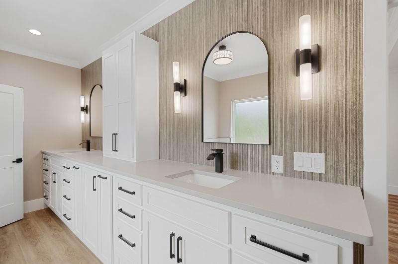 Modern bathroom with white cabinets, a long countertop, arched mirror, and vertical sconces.