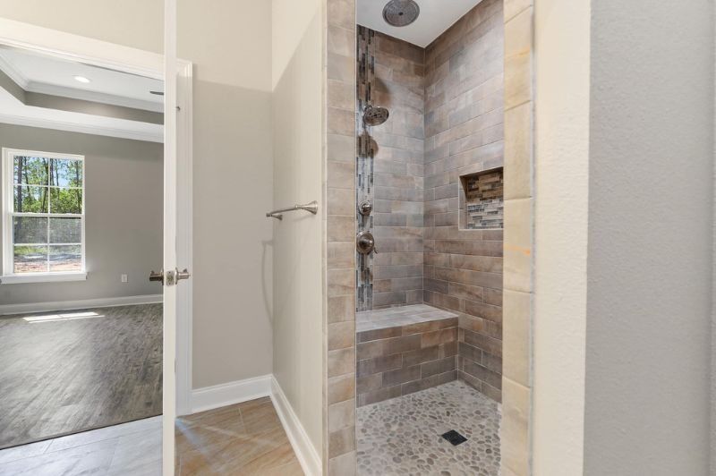 Bathroom with a tiled shower and a bench, open doorway to a bedroom with a window.