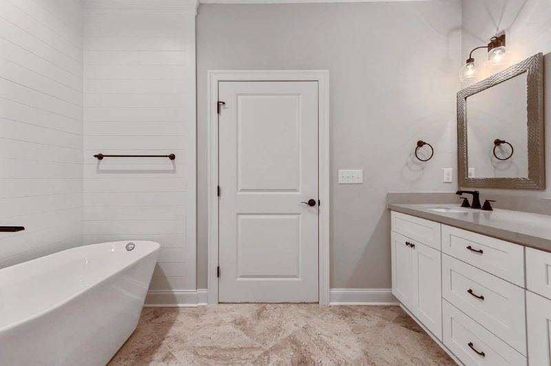 Bright bathroom with white cabinets, tub, and door. Neutral tones, vanity with mirror, black hardware.