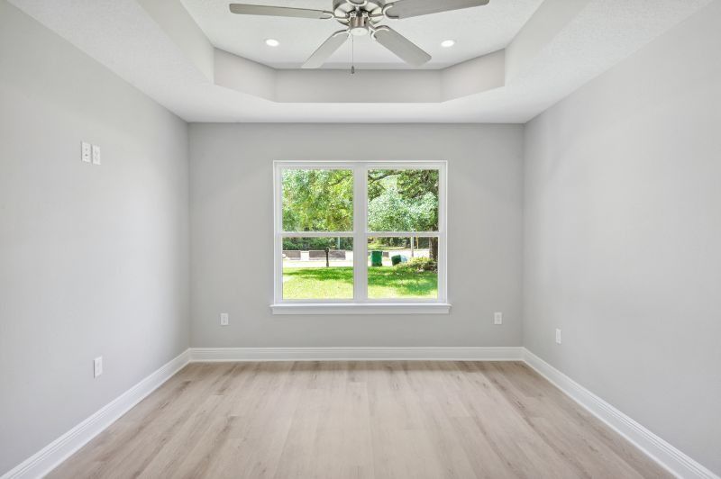 Empty bedroom with gray walls, light wood floor, window overlooking green space. Ceiling fan.