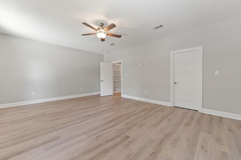 Empty, light-filled bedroom with light wood flooring, gray walls, white doors, and a ceiling fan.