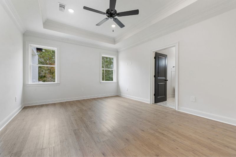 Empty, bright bedroom with two windows, a dark door, and light wood flooring.