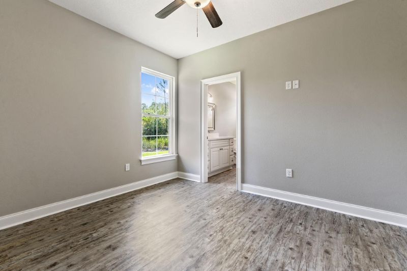 Empty bedroom with gray walls, window, open bathroom door, and wood-look flooring.