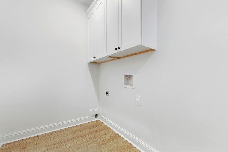 Empty laundry room with white cabinets, light wood floor, and utility hookups.
