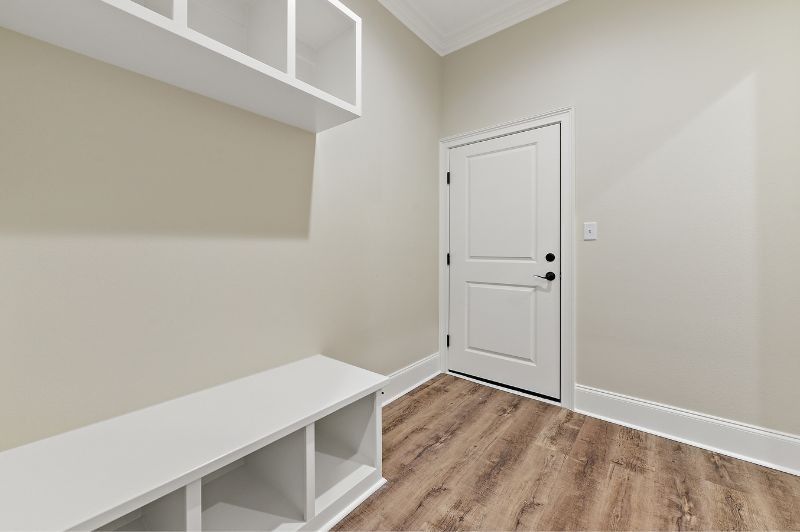 A small entryway with a white bench, shelves, and a door; beige walls and wood-look flooring.