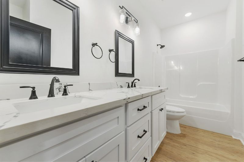 Bathroom with white cabinets, marble countertop, black fixtures and mirrors, and wood floor.