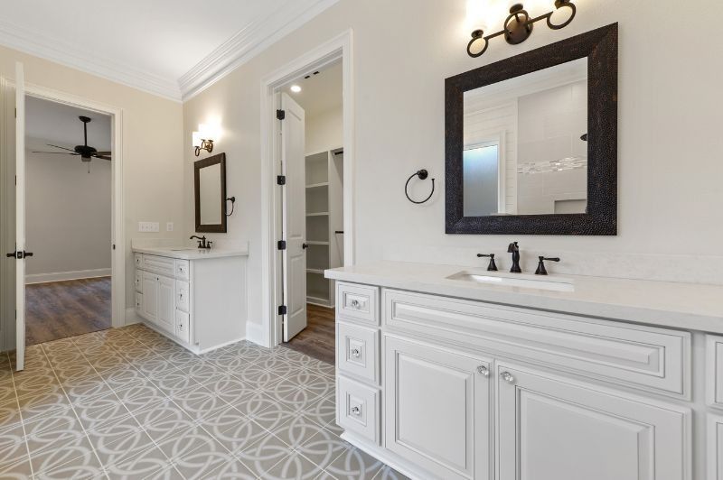 Spacious bathroom with white cabinets, patterned tile, and a dark framed mirror. Closet and another room visible.