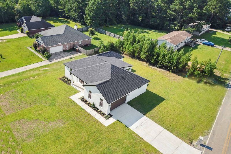 Aerial view of a modern house with a black roof and long driveway, surrounded by green lawns and other homes.