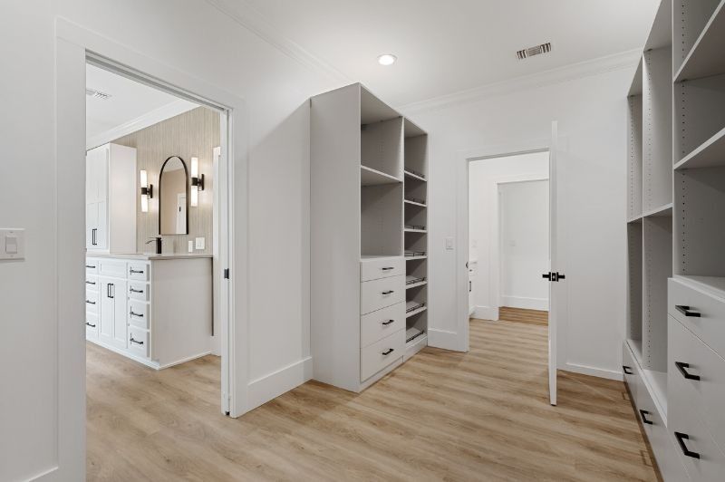 Walk-in closet with white cabinets, shelving, and light wood floors, leading to a bathroom with a vanity.