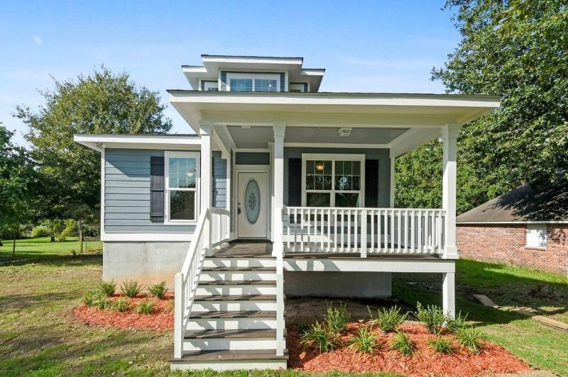 Small blue house with white porch, stairs, and trim; surrounded by green grass and landscaping.