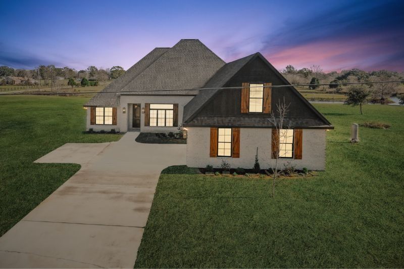 A modern, two-story house with a long driveway, set on a large grassy lawn under a twilight sky.
