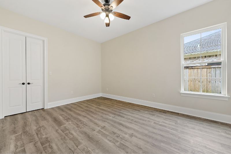 Empty bedroom with wood-look flooring, closed white double doors, window, and beige walls. Ceiling fan.