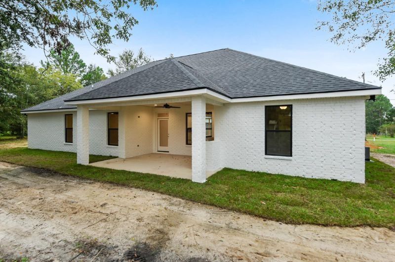 White brick house with dark roof and porch, set on grassy land, overcast sky.