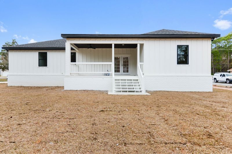 White house exterior with a porch and steps leading to a door, on a brown dirt lot, under a blue sky.