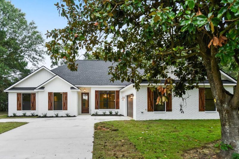 White brick house with a dark roof, brown shutters, and a curved driveway, tree in the foreground.