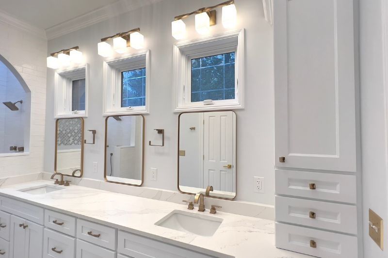 White bathroom with three mirrors, sinks, and cabinets; lit by overhead lights and a window.