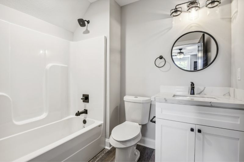 White bathroom with tub/shower, vanity, and toilet; black fixtures, round mirror.