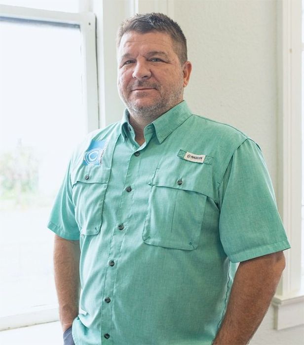 Man in a green shirt smiles, standing near a window.
